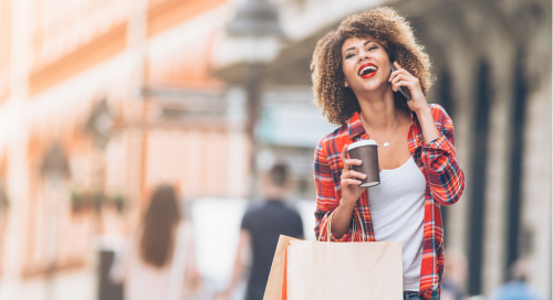 A woman laughing while talking on a phone with coffee cup and shopping bags in hand.